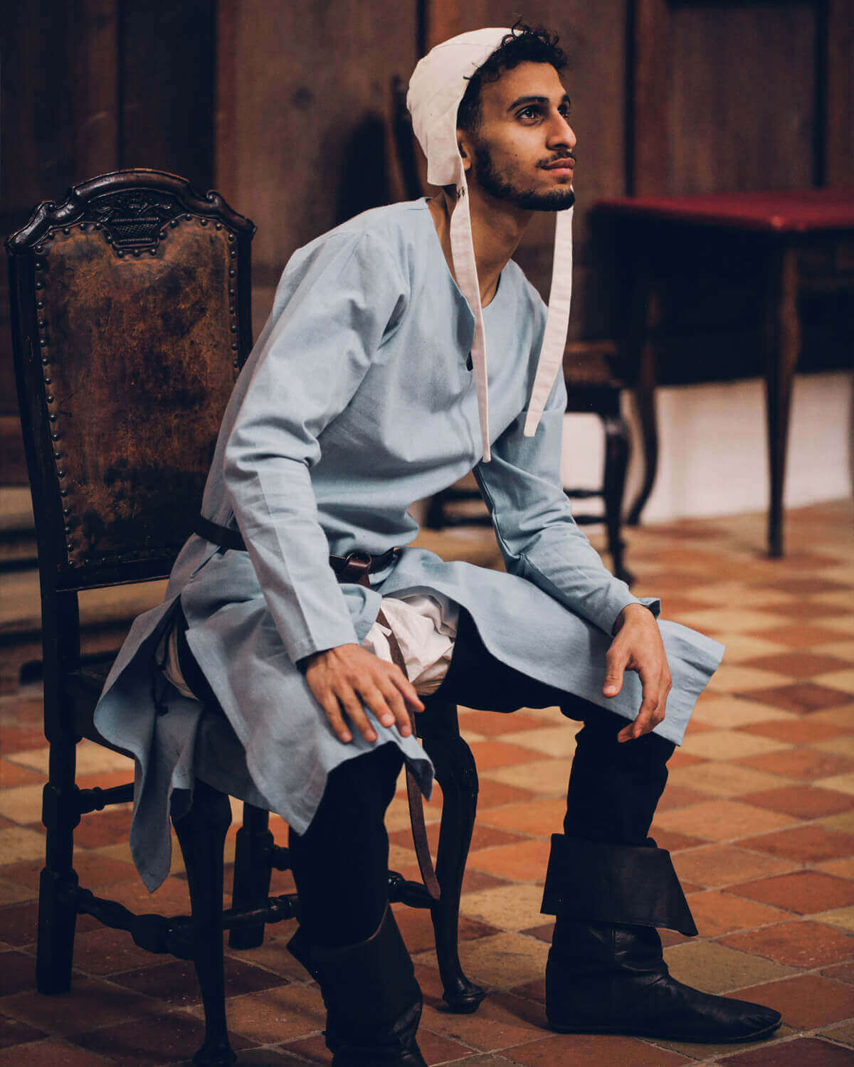Man in riding tunic, coif, braies, and hose sitting on a medieval chair.