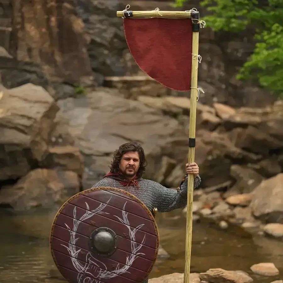 Medieval warrior holding a shield and banner by a river with rocky background, showcasing historical attire.
