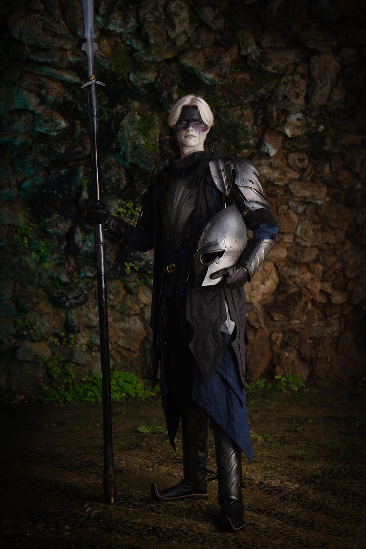 Person in dark costume holding a sword and helmet against a textured wall.
