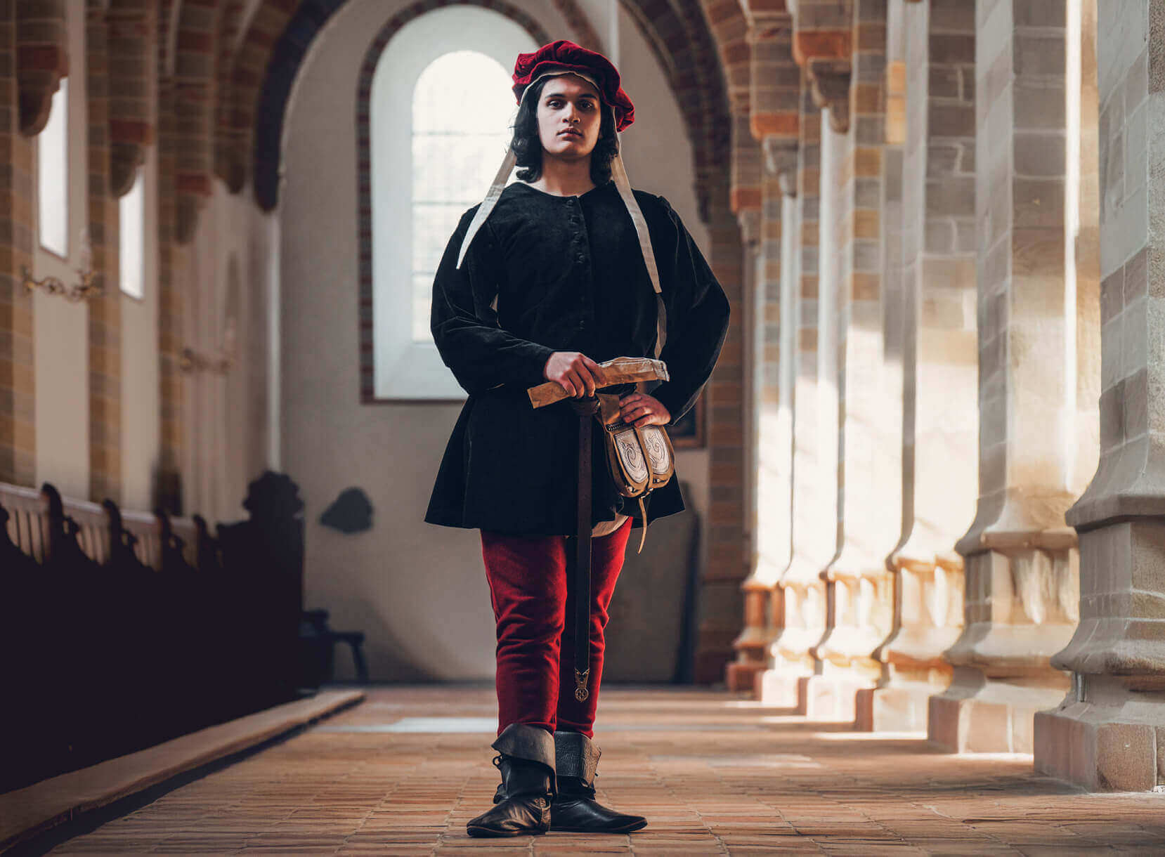 Man in Late Medieval attire with red hat and tunic, holding a dagger.