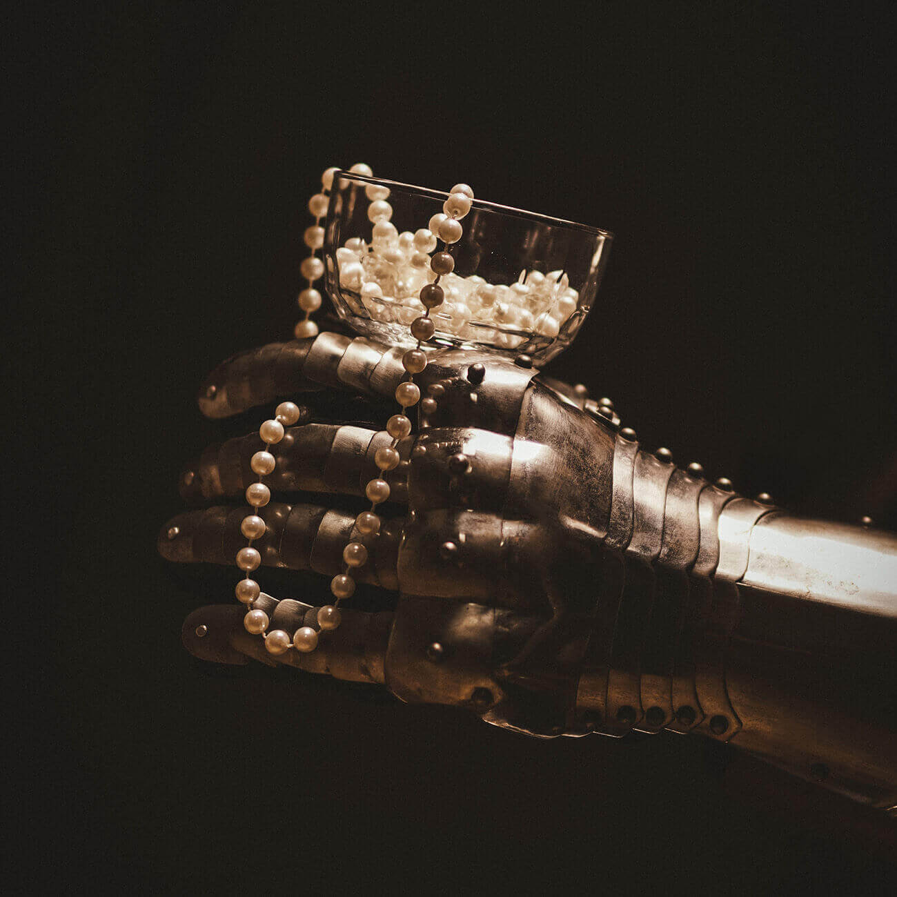 A metallic robotic hand holding a glass bowl filled with pearls and a pearl necklace draped nearby.