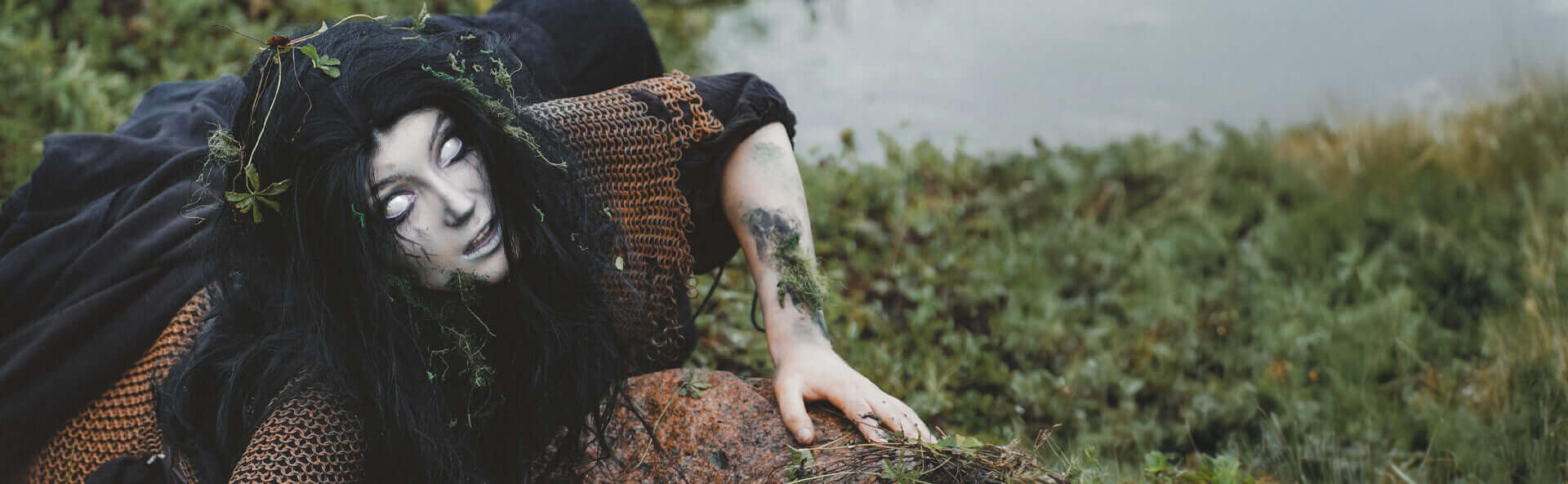 Person in dark costume with leaves, lying on grass by a body of water