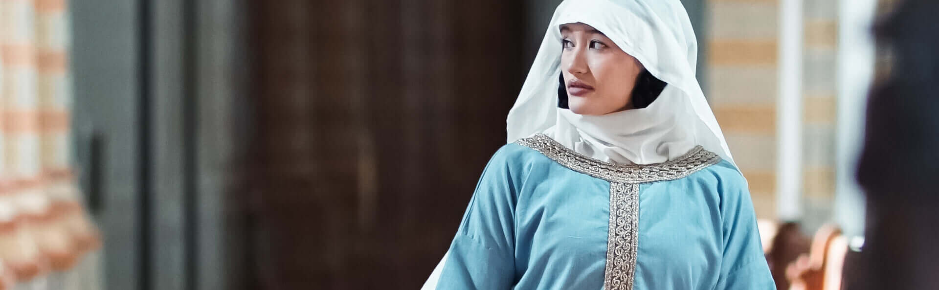 Woman wearing a light blue dress with intricate patterns and a white headscarf in an indoor setting.