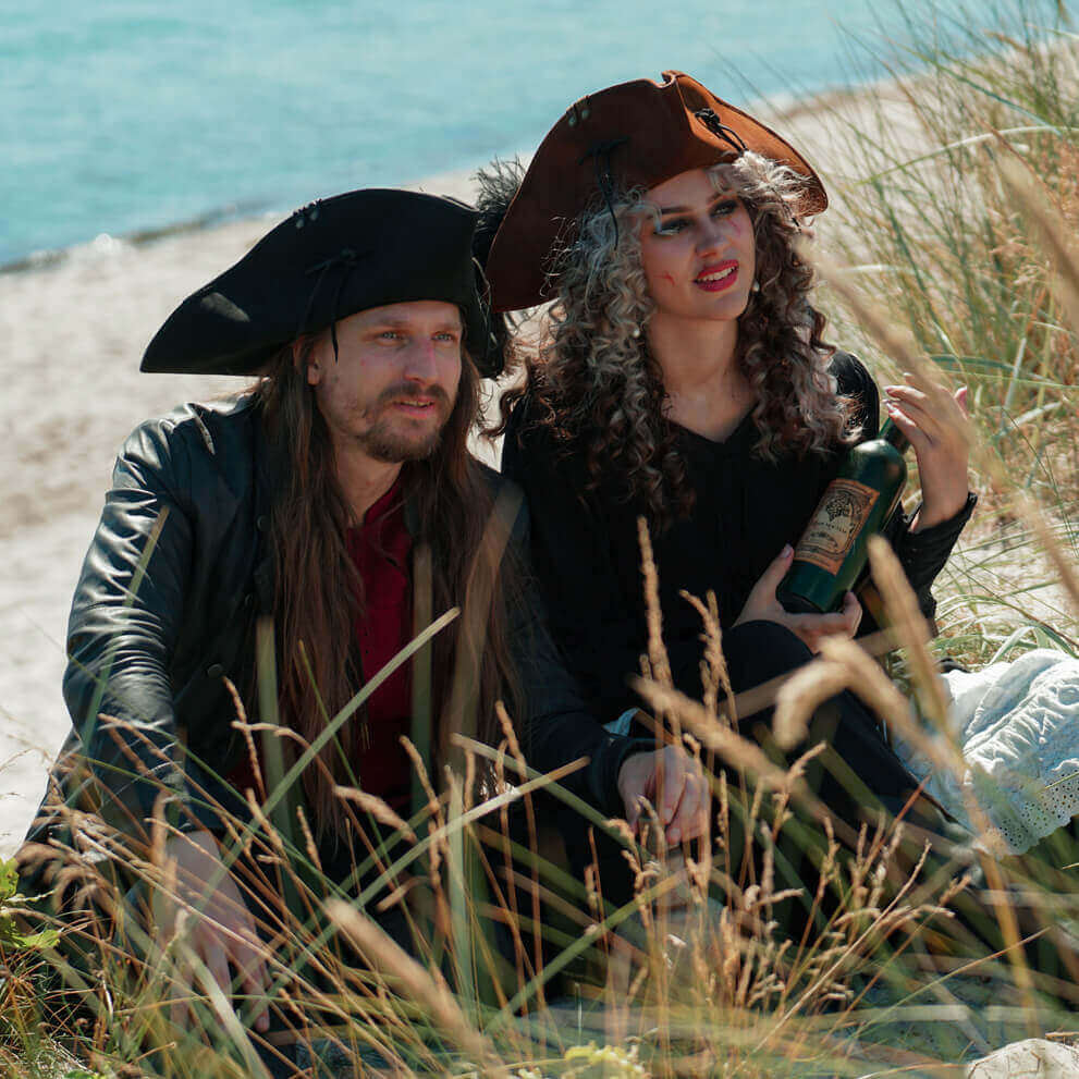 Two people in pirate costumes sitting on a beach with grass and water in the background.
