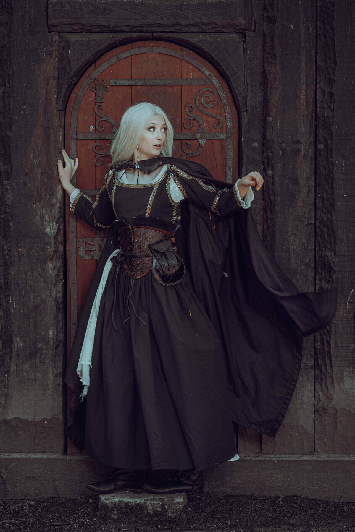 Person in dark medieval-style dress standing in front of a wooden door with intricate carvings.
